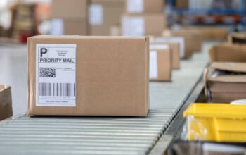 A brown cardboard box sits on a conveyor belt in a distribution warehouse. It has a priority mail label on it as well as a bar code and QR code.
